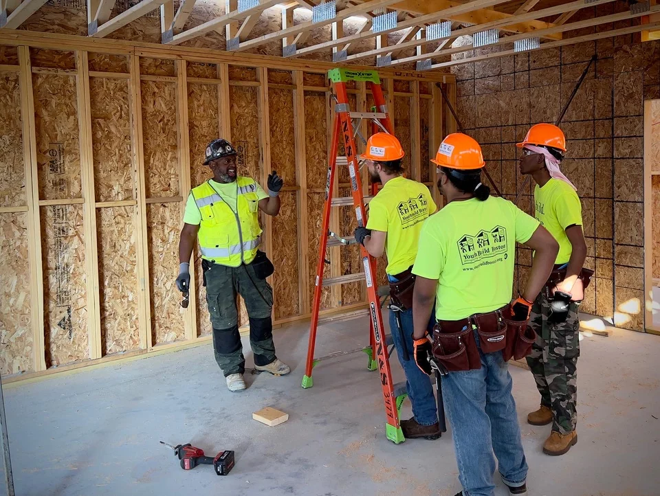 Four workers in high-visibility shirts, hard hats and tool belts standing beside an orange step ladder inside a wood-framed building while one gestures during an on-site discussion